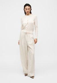 Γυναίκα που φοράει μπεζ ζακέτα με κουμπιά, φαρδιά εκρού παντελόνια λινά και παπούτσια με λεοπάρ μυτερό δάχτυλο, στέκεται μπροστά σε ένα απλό φόντο.