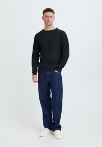 Zwarte gebreide trui met een ronde hals, gecombineerd met losse donkere spijkerbroeken en witte sneakers, tegen een lichte achtergrond.