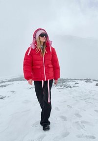 Jasnoczerwona puchowa kurtka z dodatkowym różowym kapturem i sznureczkami, noszona na jasnej bluzce i czarnych spodniach, na tle zaśnieżonego krajobrazu.