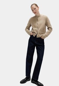 Beige stickad cardigan med ribbade muddar och fåll, med två framfickor och knappar i silverton. Kombinerad med mörkblå jeans med raka ben och svarta loafers.