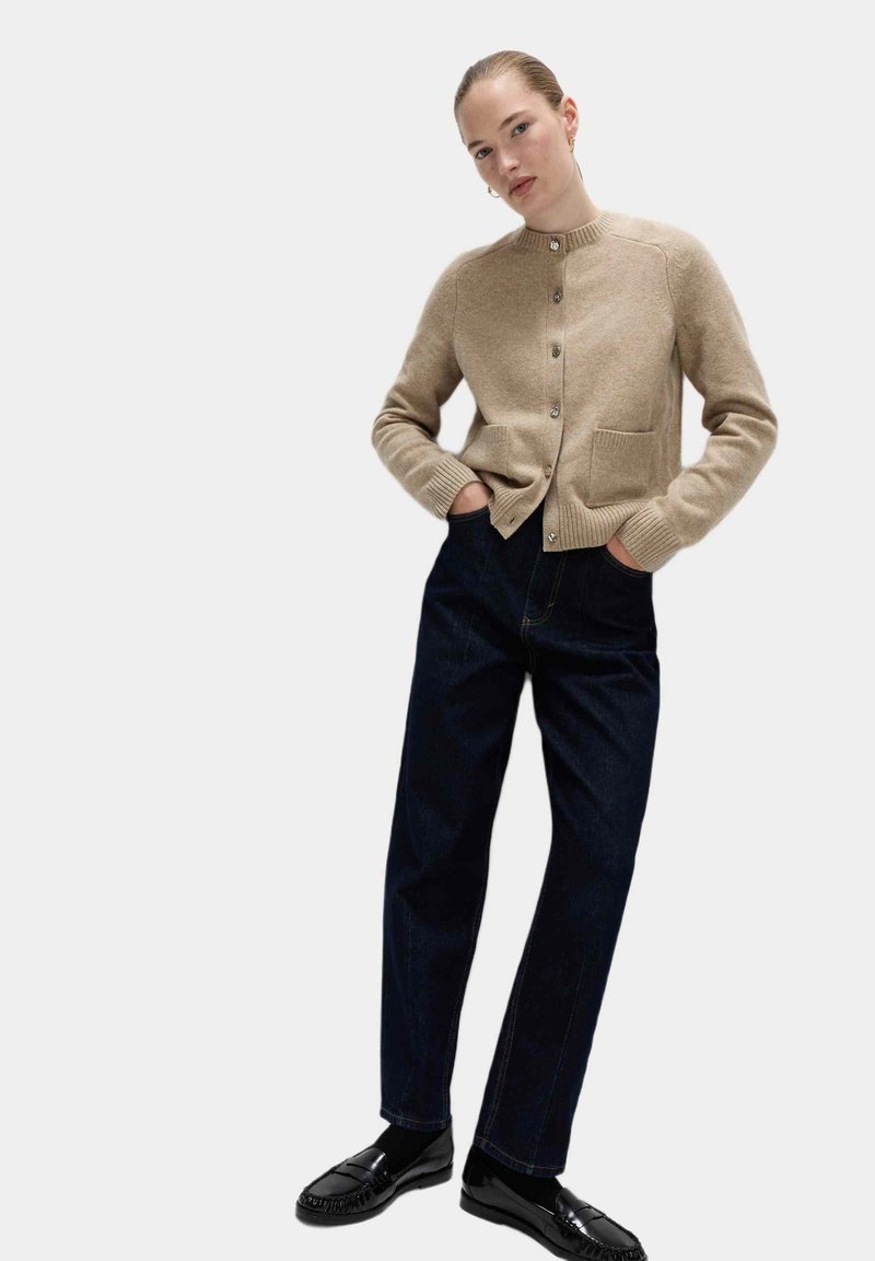 Beige stickad cardigan med ribbade muddar och fåll, med två framfickor och knappar i silverton. Kombinerad med mörkblå jeans med raka ben och svarta loafers.