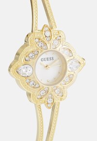 Goldwatch with floral design; embellished with clear crystals. Features a white face with gold hands and "GUESS" branding. Elegant double band.