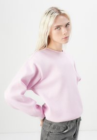 Felpa di un rosa chiaro con scollatura rotonda e design con spalle cadenti, realizzata in tessuto morbido. Abbinata a pantaloni grigi, con polsini a coste.
