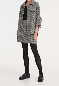 Camisa chaqueta de cuadro en blanco y negro, de gran tamaño, con dos bolsillos en el pecho, combinada con una falda a juego, medias negras y mocasines.