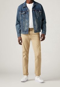 Giacca di jeans blu scuro con dettagli sbiaditi, abbinata a chinos beige e sneakers bianche. L'outfit presenta una t-shirt bianca semplice sotto.