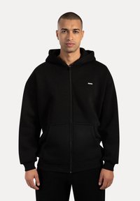 Sort zip-hoodie lavet af blød stof, med en frontlomme, ribbede ærmer og kant, samt et lille logo på brystet.