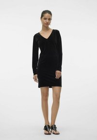 Robe longue à manches longues en velours noir avec un décolleté en V, silhouette ajustée et ourlet au genou. Accompagnée de sandales noires à bout ouvert.