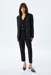 Zwarte blazer met streepjes over een aansluitend zwart gilet, gecombineerd met op maat gemaakte zwarte broeken en glanzende zwarte slingback hakken.