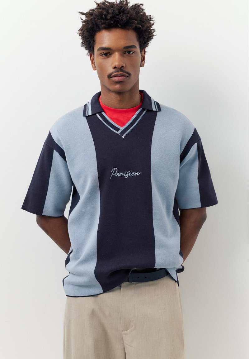 Junger Mann mit kurzen lockigen Haaren trägt ein marineblaues und hellblau gestreiftes Poloshirt mit dem Schriftzug "Parisien", beige Hosen und ein rotes Unterhemd.