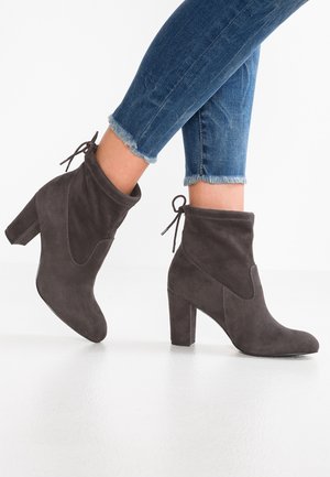 Voeten in blauwe jeans met rafelige zoom, die donkergrijze suède enkellaarzen dragen met blokhakken en een strikdetail achteraan op een witte vloer.