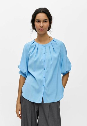 Femme debout portant un chemisier bleu clair boutonné avec manches retroussées et un pantalon large gris foncé sur un fond blanc uni.
