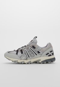 Vaaleanharmaat juoksukengät, joissa on verkko- ja synteettistä materiaalia, mustilla yksityiskohdilla, teksturoitu pohja ja merkittävä kantapään nyöri.