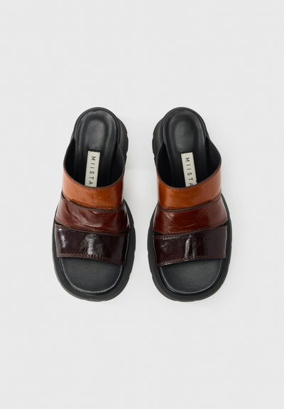 Καφέ δερμάτινα σανδάλια με μαύρες σόλες· διαθέτουν σταυρωτούς ιμάντες με ποικίλες υφές, συμπεριλαμβανομένων γυαλιστερών και ματ φινιρισμάτων, προσφέροντας μοντέρνο σχεδιασμό.