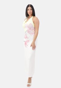 Femme aux longs cheveux noirs, portant une robe maxi sans manches jaune et blanche avec un imprimé floral rose et des chaussures à talons ouverts blanches, se tenant devant un fond blanc.