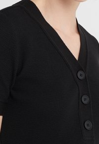 Černý pletený cardigan s texturou, s výstřihem do V a čtyřmi černými knoflíky na zapínání. Má krátké rukávy a elegantní zakončení na okrajích.