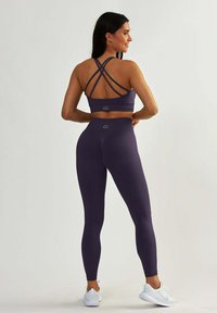 Fioletowy sportowy top crop z krzyżującymi się ramiączkami oraz pasującymi legginsami o wysokim stanie. Posiada miękki materiał i minimalistyczne detale brandingowe.