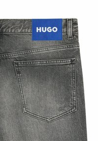 Gray denim jeans with a blue leather brand tag labeled "HUGO." Features a back pocket with stitching detail and a smooth texture.