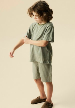 Dziecko z kręconymi włosami, ubrane w zielony koszulkę w paski z krótkim rękawem i pasujące szorty, poprawiające rękaw, stojące na beżowym tle w brązowych kapciach.