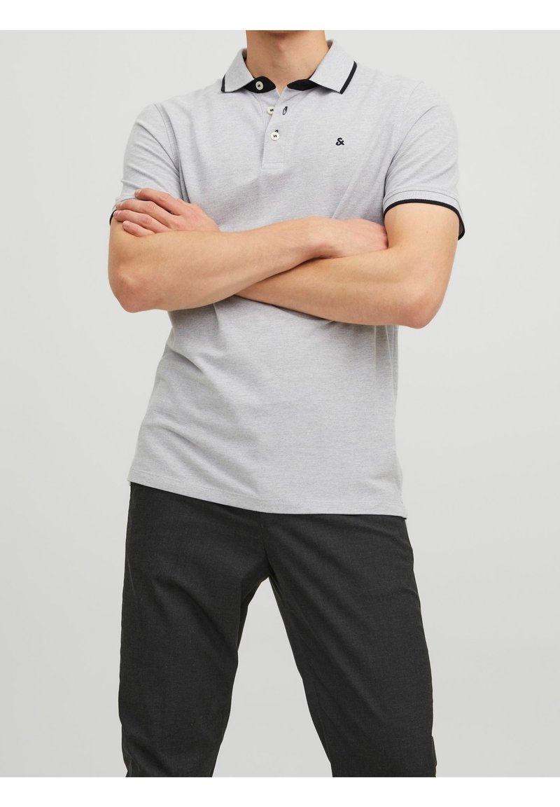 Homme portant un polo gris clair avec des bordures noires et un pantalon foncé, debout les bras croisés devant un fond uni.