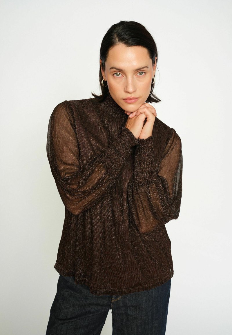 Ruda permatoma palaidinė su aukštu apykaklės kraštu, tekstūruotu audiniu, susirinkusiomis rankovėmis ir elastingais riešo rankogaliais, derinama su tamsiais džinsais.