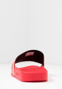 Röd halksandaler med en strukturerad sula, slät ovansida och tre vita ränder på sidan. Inredningen har en vadderad svart foder.