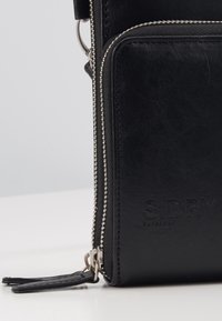 Musta nahkainen olkalaukku, jossa on vetoketjullinen tasku, hopeiset yksityiskohdat ja kohokuvioitu logo. Sujuva pinta ja tyylikäs muotoilu.