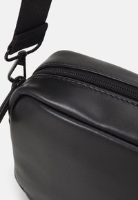 Černá syntetická kožená crossbody taška s hladkým povrchem, zipovým uzávěrem a černými kovovými doplňky. Popruh je nastavitelný s klipem.