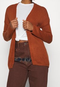 Cardigan marron en tricot avec une texture côtelée, ouverture devant, et manches longues, porté sur un haut blanc et associé à un pantalon marron.
