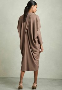 Lång, löst sittande klänning i mjukt brunt tyg, med en draperad design och en slät textur. Baksidan har en veckdetalj.
