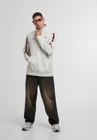 Felpa Nike grigia chiara con logo nero, caratterizzata da dettagli a strisce rosse e nere. Abbinata a jeans neri larghi e scarpe da ginnastica multicolori.