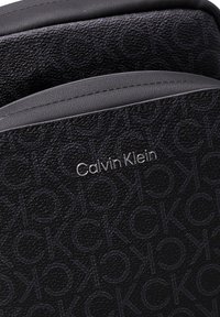 Borsa nera in tessuto con un motivo a logo ripetuto e un'etichetta argentata "Calvin Klein". Superficie liscia con bordi angolati.