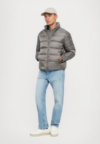 Bărbat stând cu mâinile în buzunare, purtând o jachetă puffer gri cu model, blugi ușor albaștri, adidași albi, o șapcă bej și un tricou polo gri.