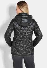 Veste doudoune noire matelassée avec une capuche, dotée d'une finition brillante, d'un motif en losange et de poignets élastiques. Portée avec un jean sombre.