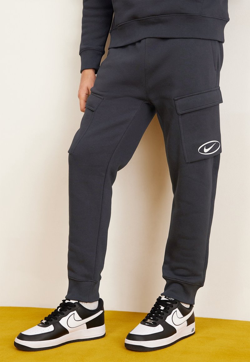 Mörkgrå sweatpants med sidofickor och en Nike-logga på låret, kombinerat med svarta och vita sneakers. Mjuk tyg, elastiska muddar.
