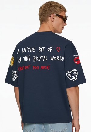 Jeune homme portant des lunettes de soleil et une chemise bleu marine avec des phrases et des patchs graphiques dans le dos et sur les manches, comprenant un soleil et des cœurs en pleurs.