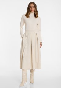 Cream turtleneck long-sleeve top paired with a pleated, long beige skirt made of ribbed fabric; knee-high beige boots complete the outfit.