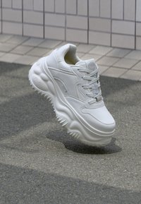 Vita klumpiga sneakers i slätt läder, med texturerade accenter, rund sula och snörningsdesign, placerade framför en grå asfaltbakgrund.