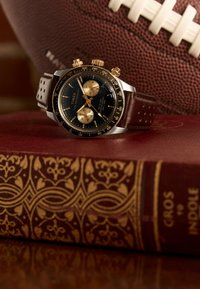 Montre Timex avec un cadran noir, des accents dorés et des fonctionnalités de chronographe ; bracelet en cuir perforé marron ; posée sur un livre rouge près d'un ballon de football.