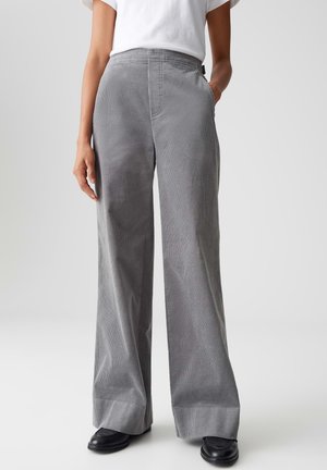 Pantalon large en velours côtelé gris avec une surface texturée, deux poches latérales et une taille haute, associé à un t-shirt blanc uni et des chaussures noires.