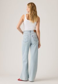 Γυναίκα με μακριά ξανθά μαλλιά που φοράει λευκό αμάνικο τοπ, ανοιχτό μπλε ψηλόμεσα παντελόνια με φαρδιά μπατζάκια και κόκκινα μυτερά παπούτσια, γυρισμένη με την πλάτη.