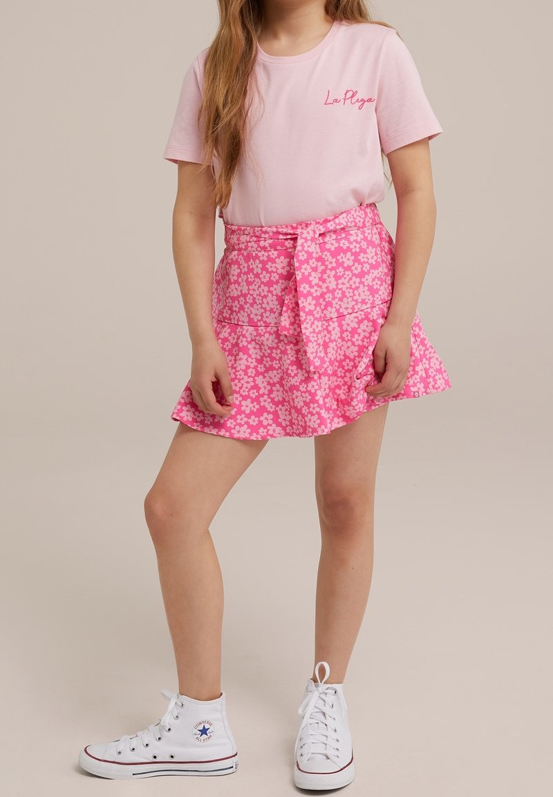 Fille portant un t-shirt rose à manches courtes avec le texte « La Playa » et une jupe fleurie rose nouée à la taille, associée à des baskets blanches montantes.