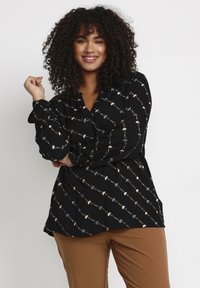Black blouse featuring a patterned design with small floral accents in blue, white, and beige. Long sleeves with gathered cuffs.