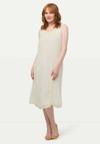 Vestido sin mangas color crema con intrincados bordados en forma de flores doradas, de longitud hasta la rodilla, confeccionado en una tela ligera, con un dobladillo en forma de ola, combinado con sandalias amarillas de tiras.