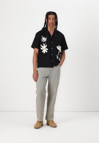 Zwart shirt met korte mouwen en witte bloemenpatronen, knoopsluiting, gecombineerd met lichte spijkerbroeken en tan kleurige schoenen. Eenvoudige, casual stijl.