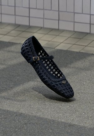 Černá tkaná plochá bota s malým páskem se sponou, vznášející se nad texturovanou betonovou podlahou a pozadím s dlaždicovou stěnou.