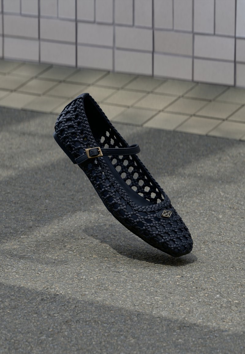 Fekete szövött lapos cipő kis csattos pánttal, amely egy texturált beton padló és csempézett fal háttere felett lebeg.