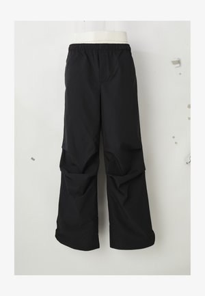 Černé volné kalhoty s elastickým pasem, zavěšené na jednoduchém světlém pozadí.