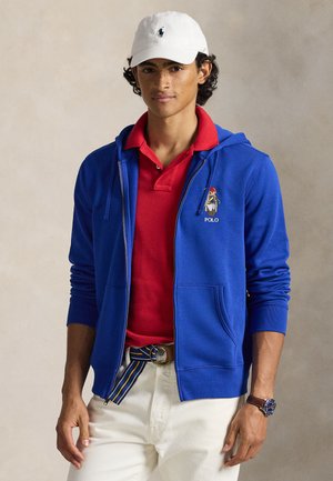 Royal blå lynlås-hoodie, rød polo-shirt under, hvid kasket, lyse bukser med lomme og logodetalje på hoodien.