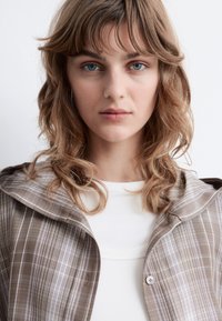 Brown plaid jacket with a hood, featuring light blue accents and a button closure, worn over a white ribbed shirt. Smooth fabric texture.