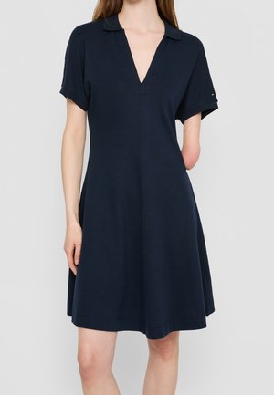 Vrouw die een marineblauwe jurk met korte mouwen en een V-hals draagt, staand tegen een effen lichtgrijze achtergrond.